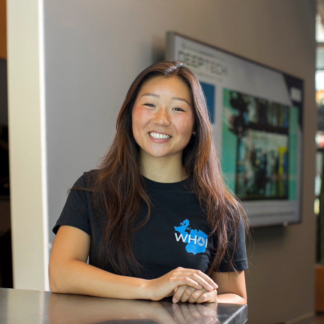 A woman photographed from the front wearing a black shirt with a Spilhaus map projection and a WHOI Acronym Logo on the front left chest. The woman is standing next to a table.