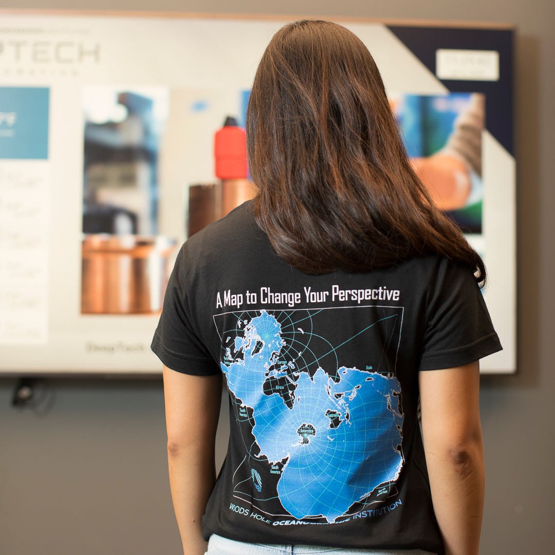 A woman viewed from behind wearing a black t-shirt with a large blue and white map graphic on the back, featuring the text 'A Map to Change Your Perspective' and 'WOODS HOLE OCEANOGRAPHIC INSTITUTION'.