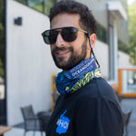 A man wearing aviator sunglasses and a blue patterned neck gaiter with 'WOODS HOLE OCEANOGRAPHY INSTITUTION' printed on it, standing outdoors in front of a modern building.
