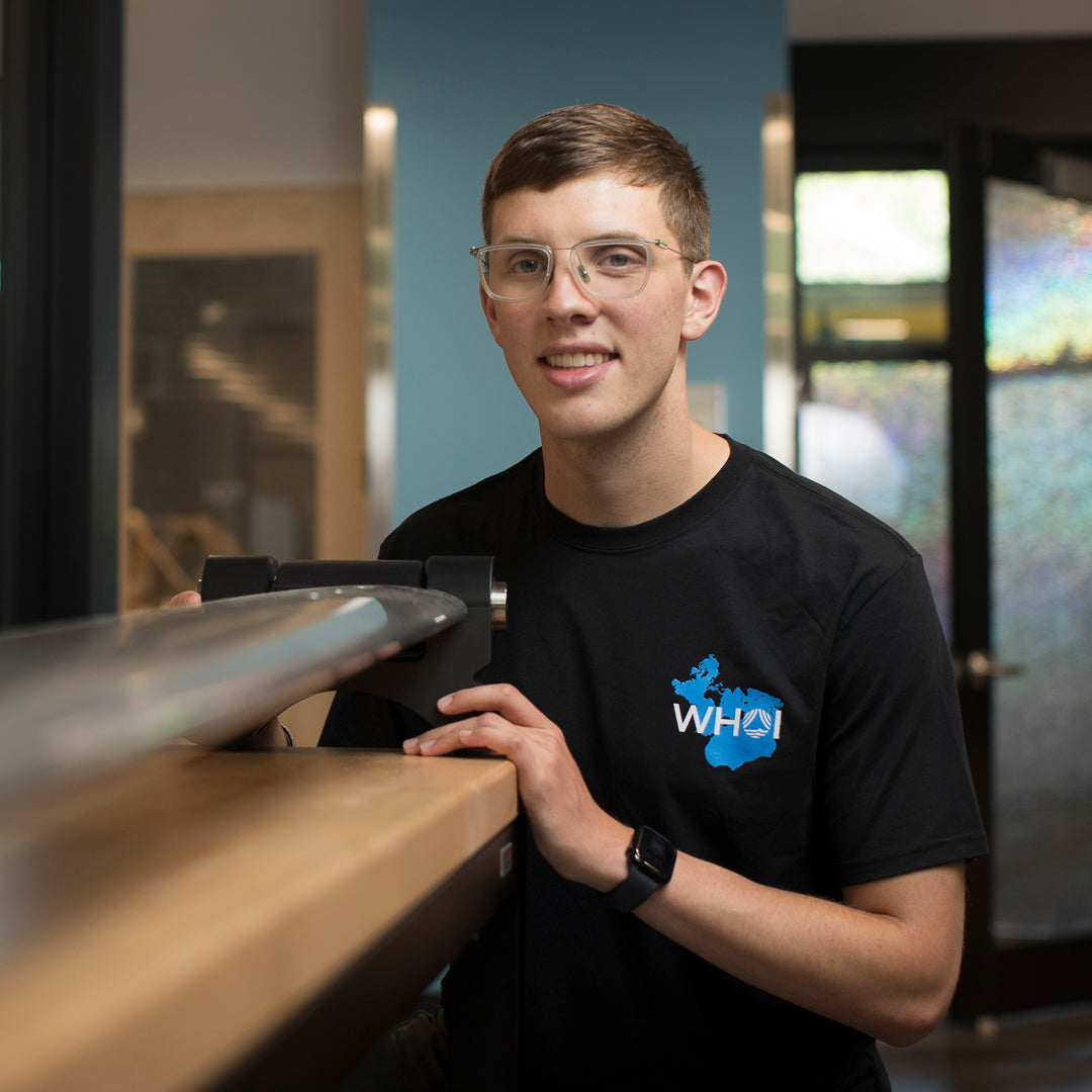 A man photographed from the front wearing a black shirt with a Spilhaus map projection and a WHOI Acronym Logo on the front left chest. Man is standing next to a large tool.