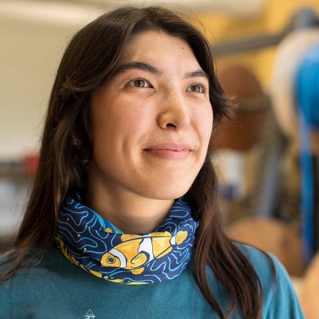 A woman wearing a neck gaiter with an orang, yellow and blue design featuring a clown fish.