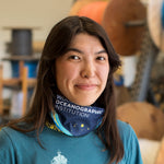 A woman with long dark hair wearing a blue bandana around her neck and a blue t-shirt, with the words 'WORLD OCEANOGRAPHIC INSTITUTION' visible on the bandana.