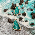 Two triangular wooden dangle earrings with a vibrant green and turquoise inlay featuring white wavy lines and dots, displayed on a textured background resembling ocean waves and dark rocks.